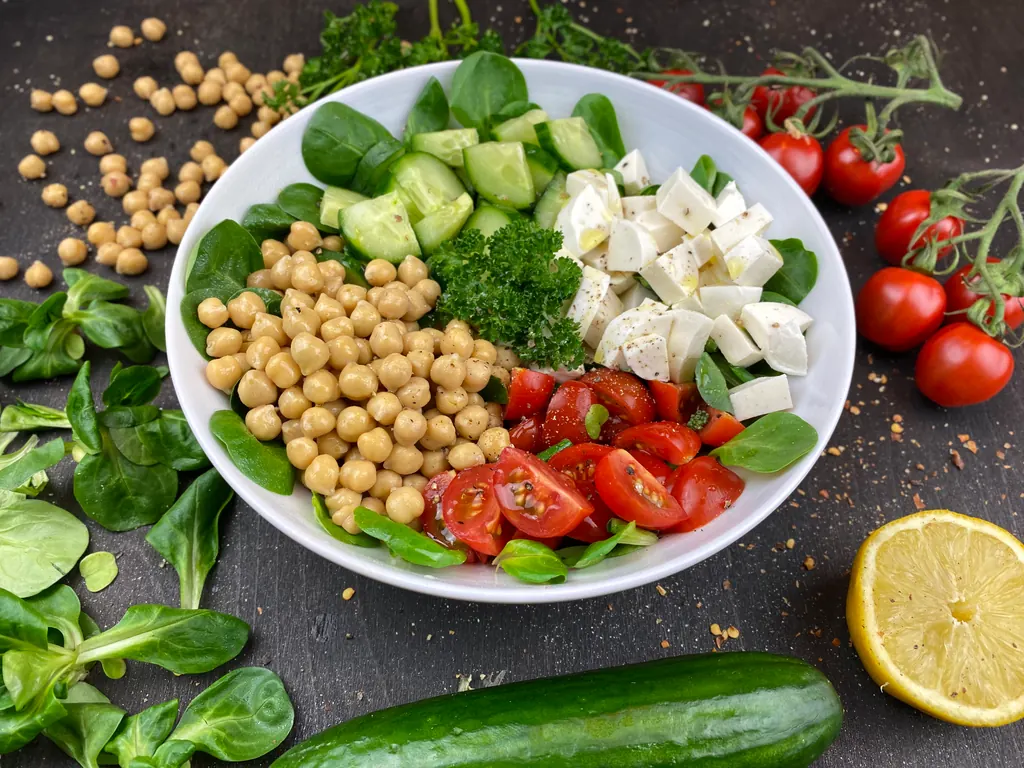 Chickpeas, cucumber, cherry tomatoes, salad leaves, parsley and white mozzarella-style cheese cubes in a bowl beside lemon.